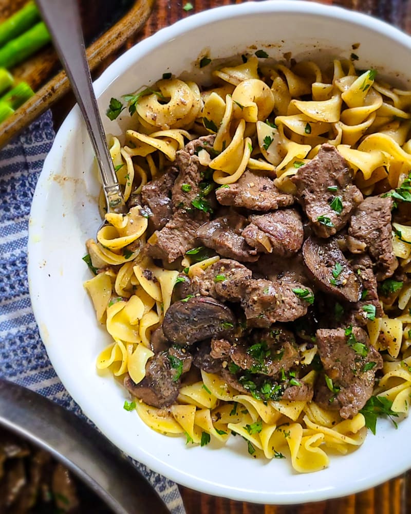Beef stroganoff over egg noodles in a white bowl, with seared beef strips, mushrooms and chopped parsley